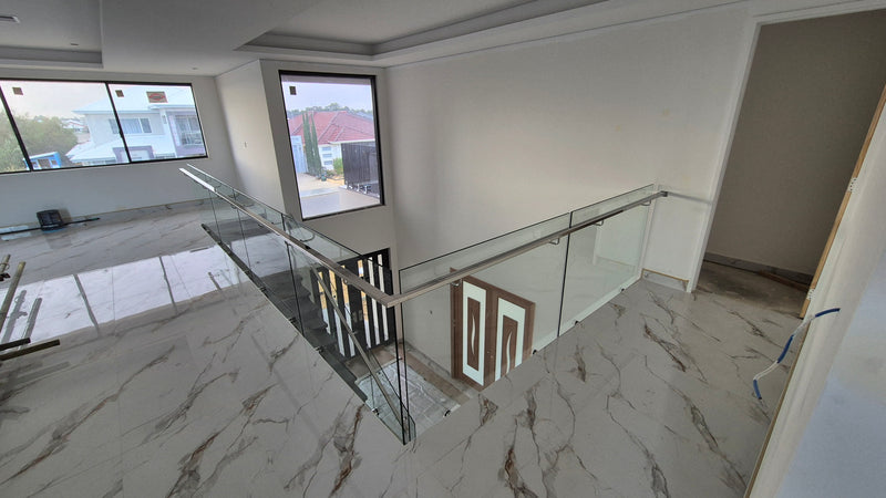 A modern glass railing with a stainless steel handrail installed on an upper-level staircase landing in a residential building with white walls and marble-patterned flooring.