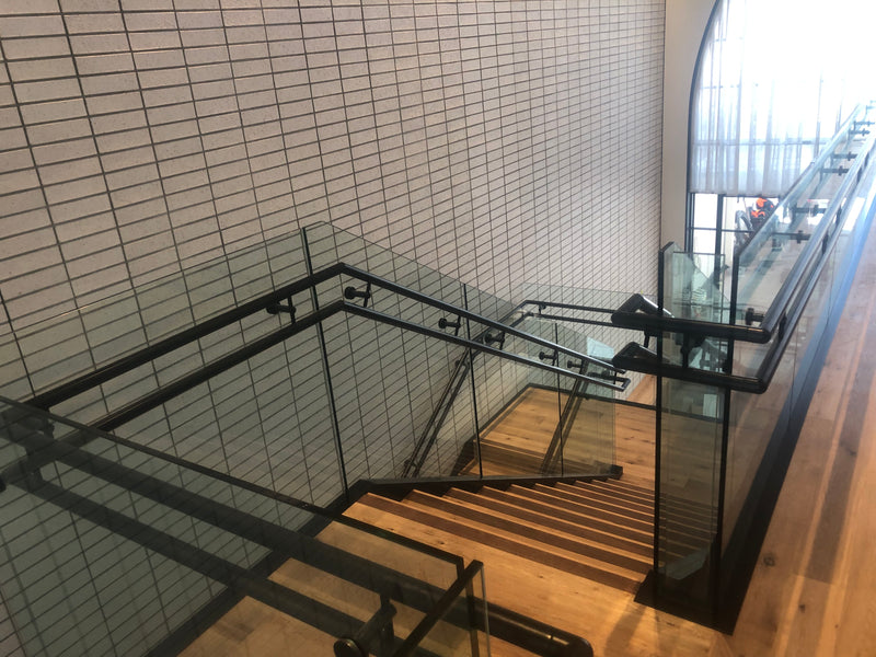 Black Base Rail project in a commercial space with double handrail on the side of the glass. The glass railing comes up a stair on both sides and then transitions to a level balcony. 