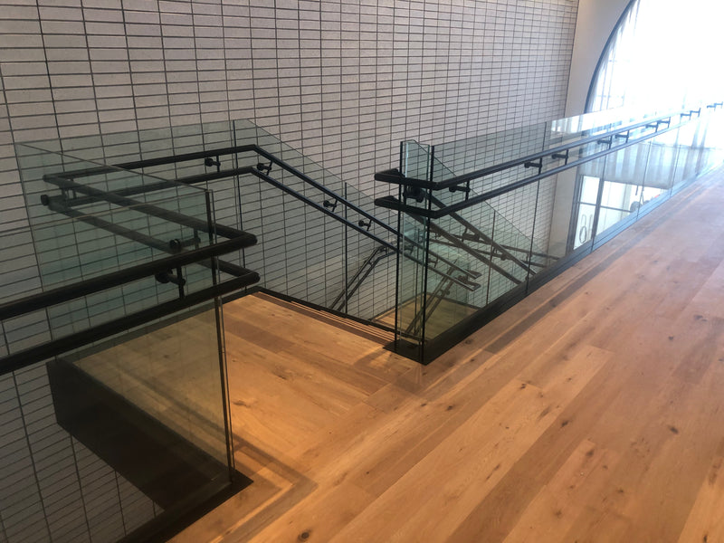 Black Base Rail project in a commercial space with double handrail on the side of the glass. The glass railing comes up a stair on both sides and then transitions to a level balcony. 