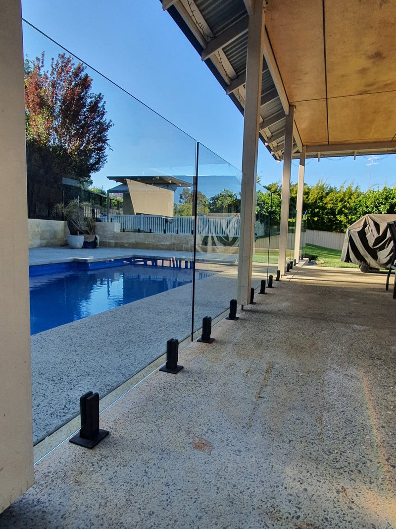 A modern glass pool fence installed along the edge of a swimming pool on a covered patio. The transparent glass panels are supported by black metal posts anchored into a speckled concrete deck, with a blue swimming pool and outdoor greenery visible in the background under a clear sky.