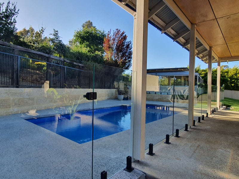 A modern outdoor swimming pool with blue water and a light concrete deck, bordered by clear glass panels supported by black posts under a covered patio area.