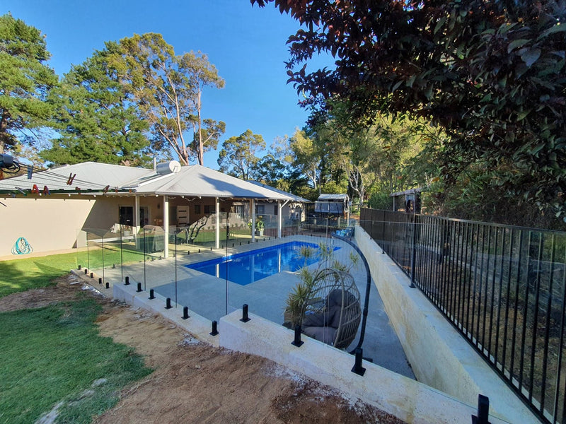 A backyard pool area with a rectangular blue swimming pool, glass panel fencing, a concrete deck, a covered patio with white canopies, grass, trees, and a clear blue sky.