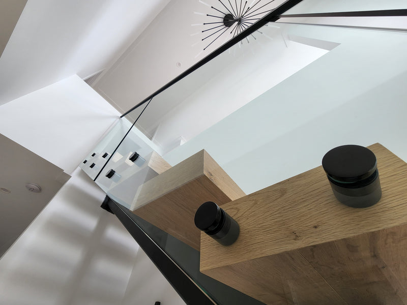 Modern interior staircase scene with light wood risers, a glass railing, black metal handrails, and two matte black cylindrical containers placed on the wooden step.