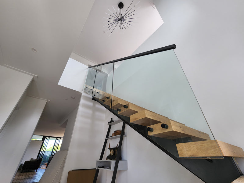 Modern floating wooden staircase with a glass railing and black metal handrails, set in a minimalist white-walled interior with a starburst ceiling light.