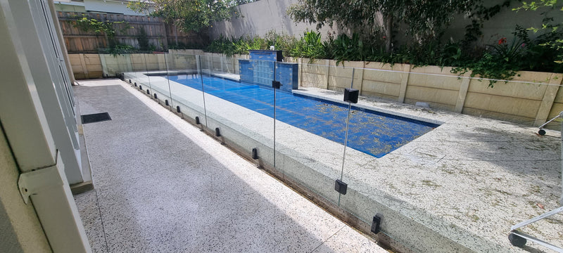 A long rectangular outdoor swimming pool with blue water and a glass safety fence, bordered by a light gray concrete deck and beige retaining walls with greenery.