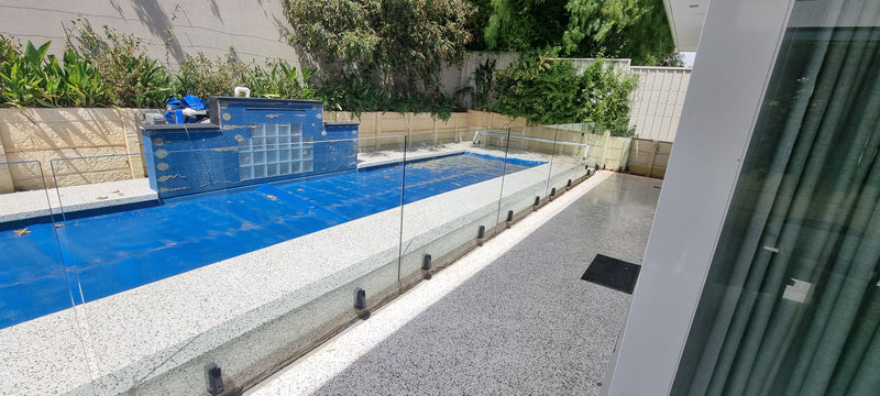 A modern outdoor swimming pool with clear glass safety railings, featuring a blue tiled end wall and a gray stone coping, set in a backyard with greenery and a house facade visible.