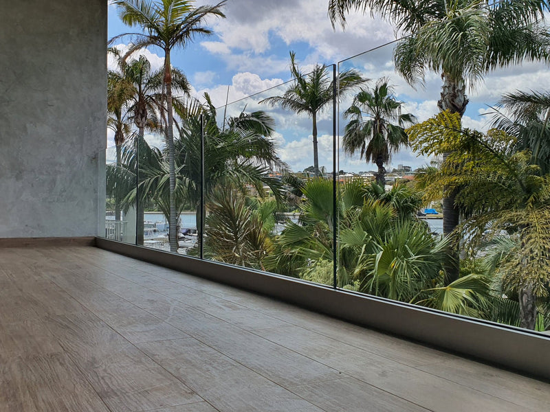 Modern glass balcony railing installed on a wooden deck, overlooking a tropical outdoor scene with palm trees and a cloudy blue sky.