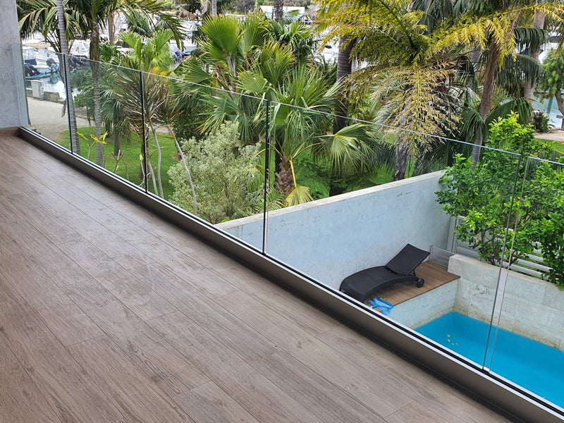 A modern interior balcony with light brown wood-look flooring and a clear glass railing, overlooking a tropical garden with palm trees, a small blue pool, and a black lounge chair.