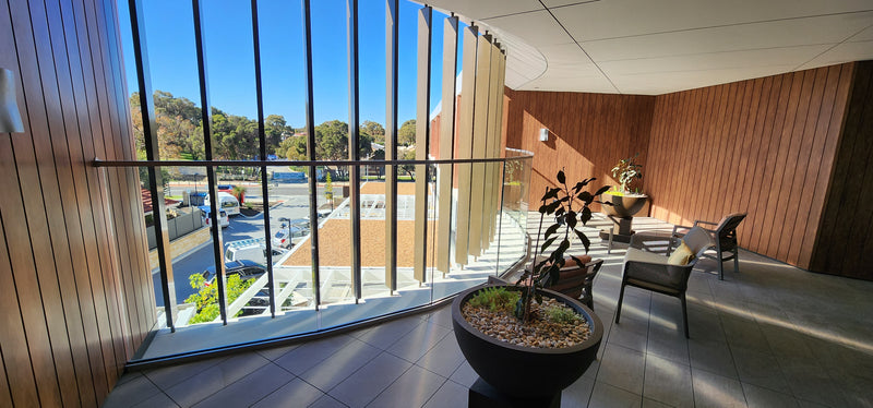 A modern outdoor patio area with wood-paneled walls, large floor-to-ceiling windows, a potted plant in the foreground, and seating visible beyond the glass overlooking a landscaped area.