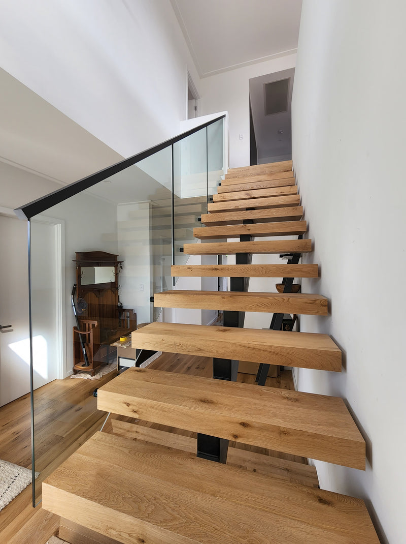 Modern floating wooden staircase with natural oak steps and a clear glass railing with a black metal handrail, set against white walls.