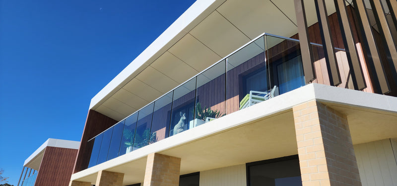 Modern glass balcony railing system installed on a multi-story residential building under a clear blue sky.