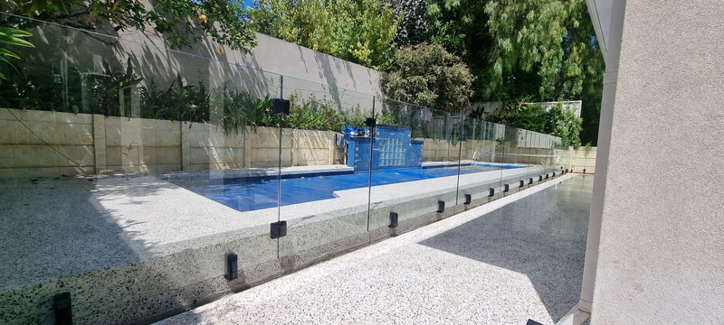 A modern outdoor swimming pool with clear glass fencing and a blue water feature, set in a landscaped backyard with trees and a stone wall.