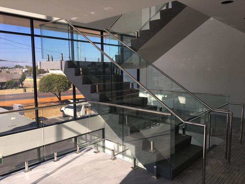 Modern staircase with glass railings and metal handrails, shown in a contemporary building interior with large windows and an outdoor view.