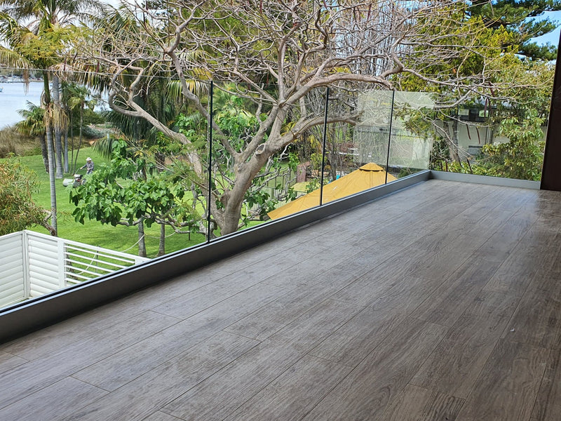 A modern wooden deck floor with a glass railing overlooking a lush green tropical garden with palm trees and a body of water in the distance.