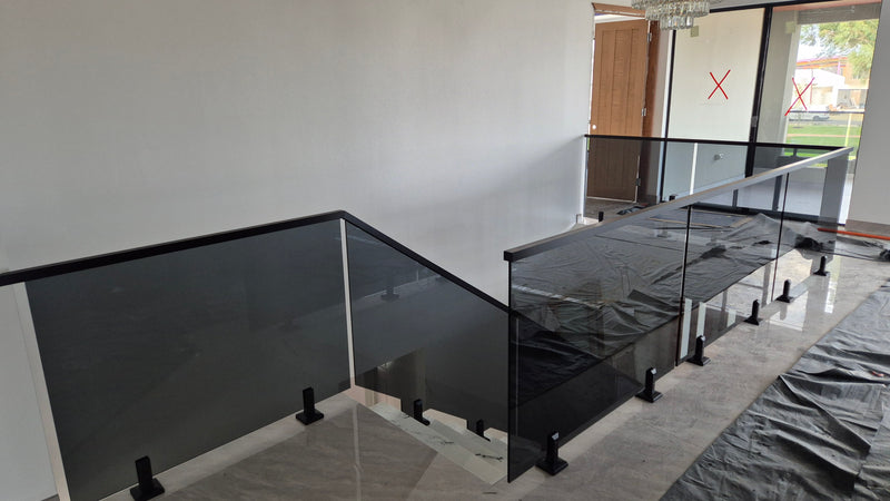 Modern glass stair railing with black metal frames and black handrails installed on an indoor staircase, with protective plastic sheeting covering parts of the railing and floor.