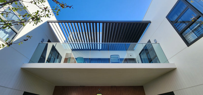 Modern white building facade with a glass-rail balcony, dark metal roof overhang, blue sky, and part of a wooden door at the bottom.