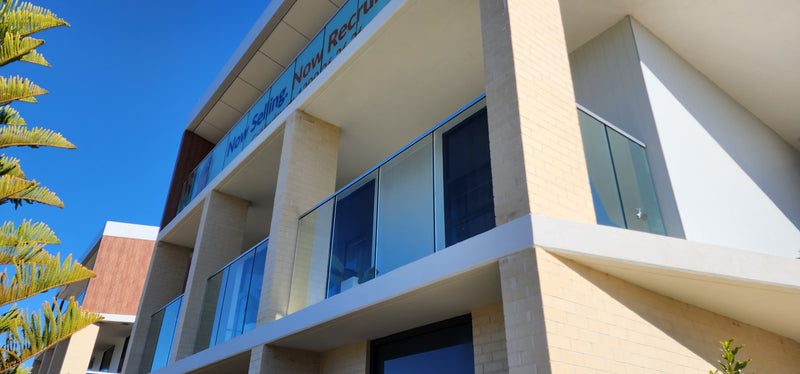 Modern multi-story building exterior with beige brick columns, glass balconies, large windows, and a blue sky background. A sign on the building reads 'Now.