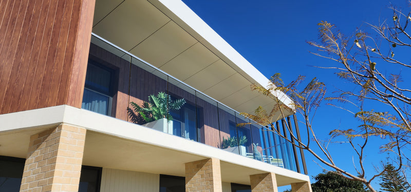 Modern two-story apartment building facade with wood paneling, glass railings, brick columns, and a clear blue sky with tree branches in the foreground.