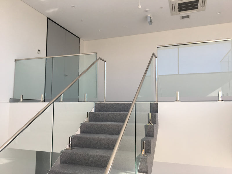 A modern staircase with gray carpeted steps and glass railings with stainless steel handrails, set in a bright, minimalist interior with white walls.