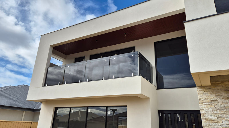 Modern two-story house exterior with a glass railing balcony, cream stucco walls, large dark windows, and a stone facade section under a blue sky with clouds.