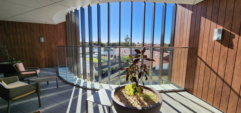 A modern outdoor balcony with wood-paneled walls, glass railings, and a large potted plant in the foreground. The balcony overlooks a residential area with houses and trees under a clear blue sky.