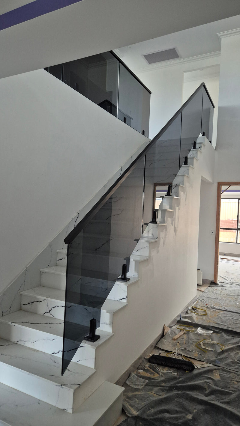 A modern staircase with white marble-look steps and a dark-tinted glass railing, shown in an interior room with white walls and a floor covered in protective sheeting.