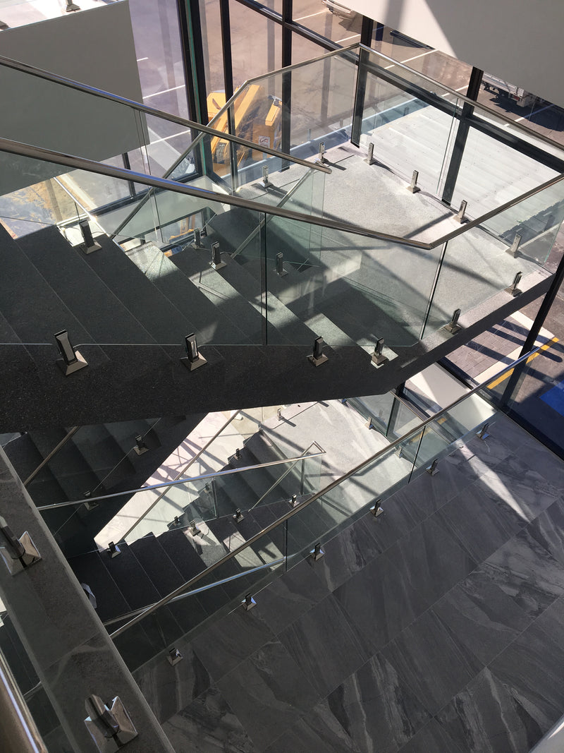 Modern glass staircase with dark gray stone steps and stainless steel handrails in a contemporary building interior.