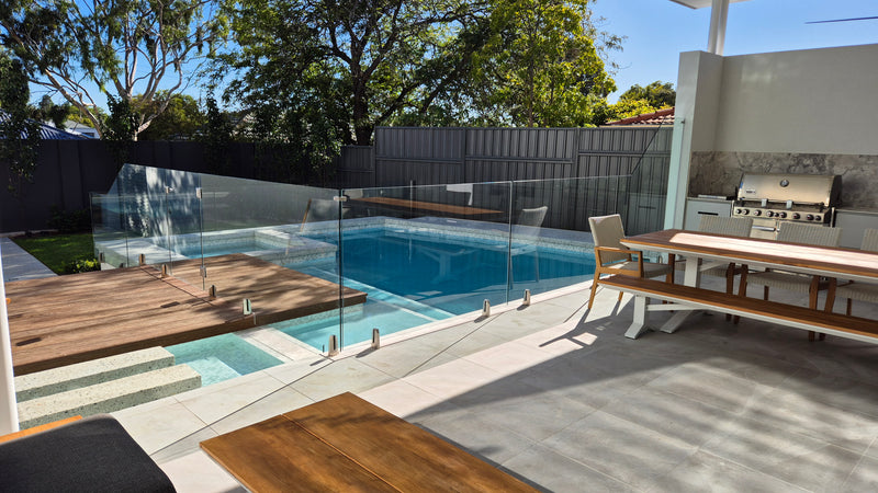 Modern outdoor backyard patio with a glass railing overlooking a blue swimming pool, a wooden deck, dining table, and built-in outdoor kitchen area with a grill.