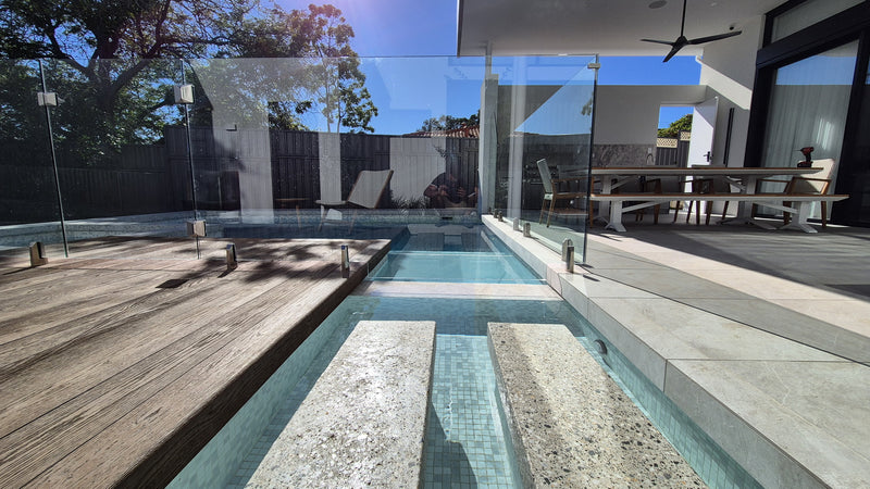 Modern outdoor plunge pool with clear glass walls, a wooden deck, stone steps, and a contemporary patio area under a covered roof.