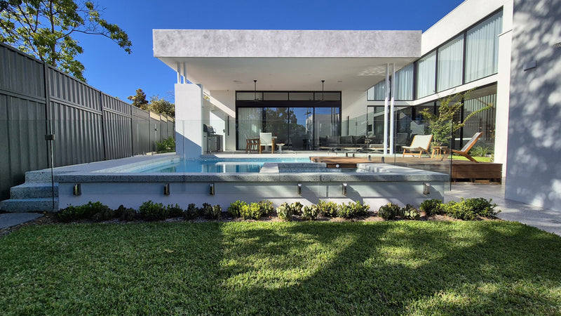 Modern luxury outdoor pool and patio area in front of a contemporary white house with large glass windows, surrounded by a manicured green lawn and trees under a clear blue sky.