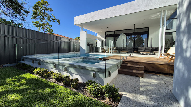 Modern outdoor hot tub and plunge pool area beside a contemporary house, with glass fencing, a wooden deck, lawn, shrubs, and blue sky in the background.