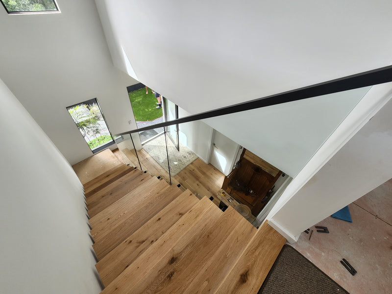 A modern staircase with light oak wood treads and a sleek black metal handrail, photographed from an elevated angle in a bright, minimalist home interior.
